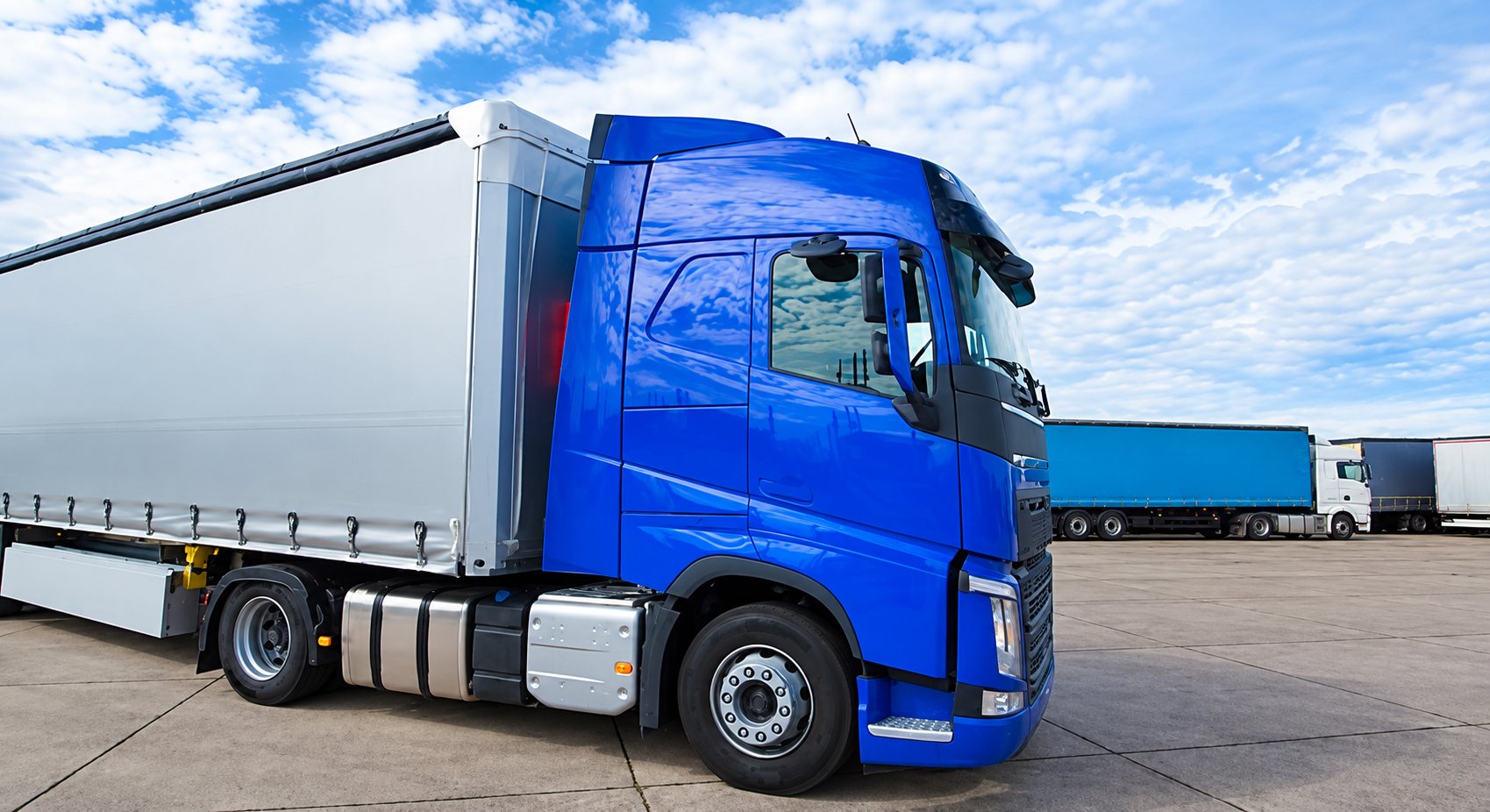 Un camion bleu attelé à une remorque grise est garé sur un grand parking, avec d'autres poids lourds en arrière-plan sous un ciel nuageux.