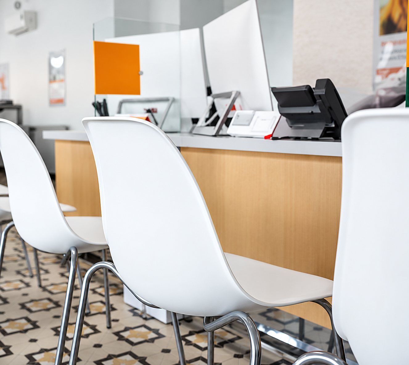 Plusieurs chaises blanches alignées devant un comptoir en bois clair avec des écrans et un terminal de paiement, sur un sol à motifs.