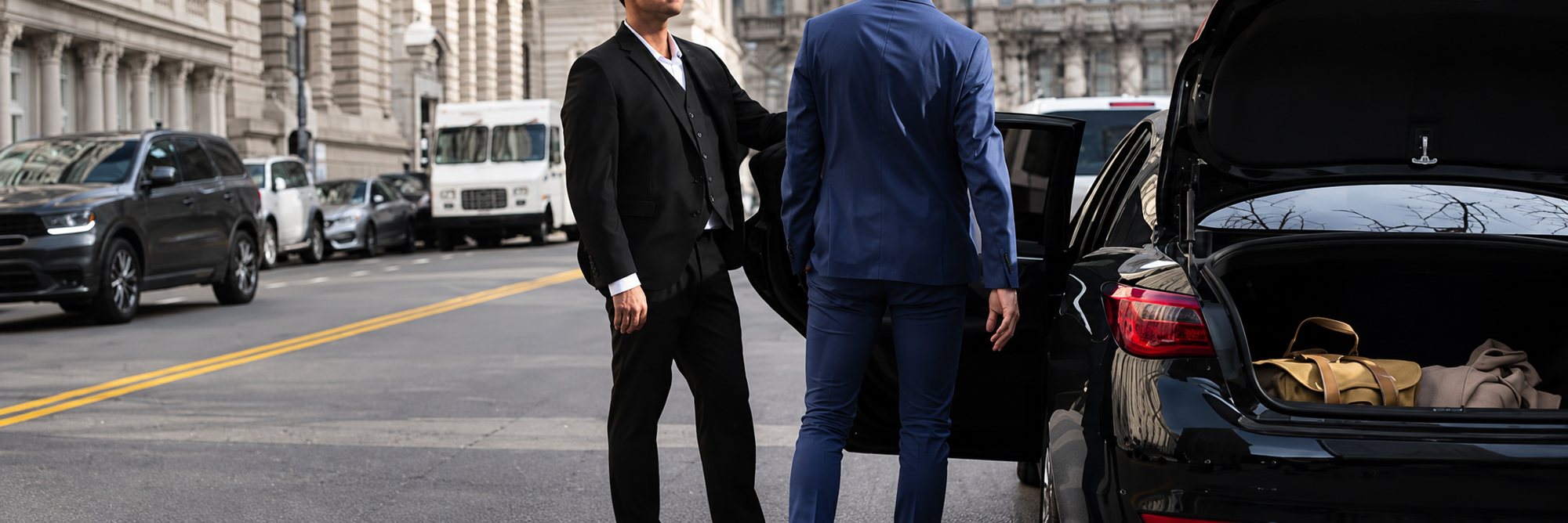 Un chauffeur en costume noir tient la portière d'une berline noire pour un client en veste bleue, coffre ouvert, dans une rue de ville.