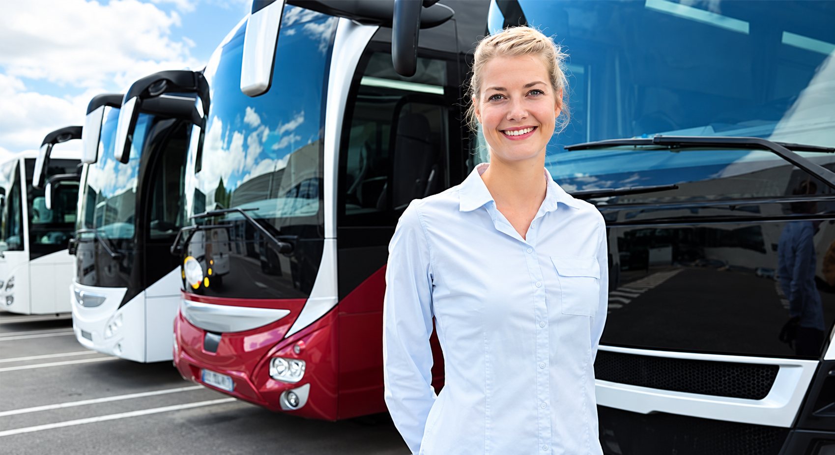 Une jeune femme blonde en chemise bleu clair sourit devant plusieurs autocars de tourisme alignés sur un parking.