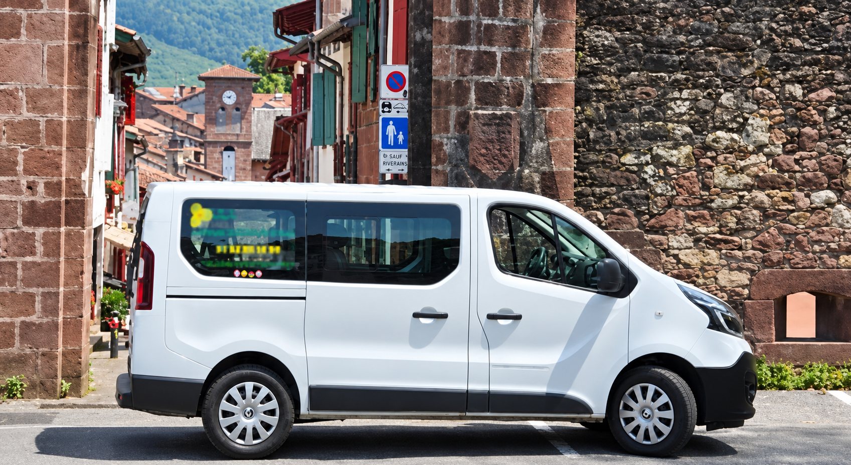 Un minibus blanc est garé dans une rue étroite d'un village entre des murs de pierre, avec des collines vertes en arrière-plan.