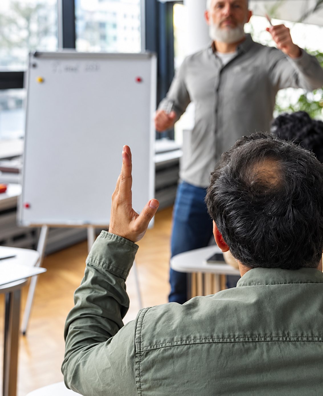 Vu de dos, un homme lève la main pendant qu'un formateur à barbe grise s'exprime devant un tableau blanc dans une salle lumineuse.