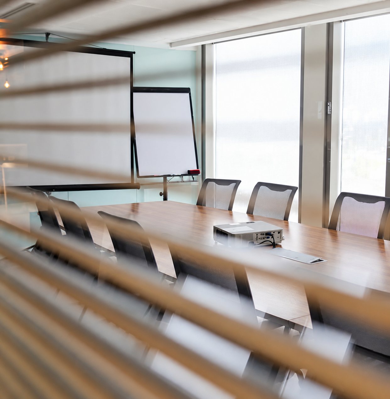 Une salle de réunion lumineuse avec une longue table en bois, un projecteur, des chaises, un écran et un paperboard, vue à travers des lamelles de stores.